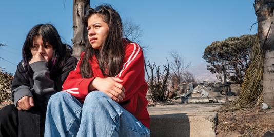 Two teens sit on curbside near wildfire destruction. 